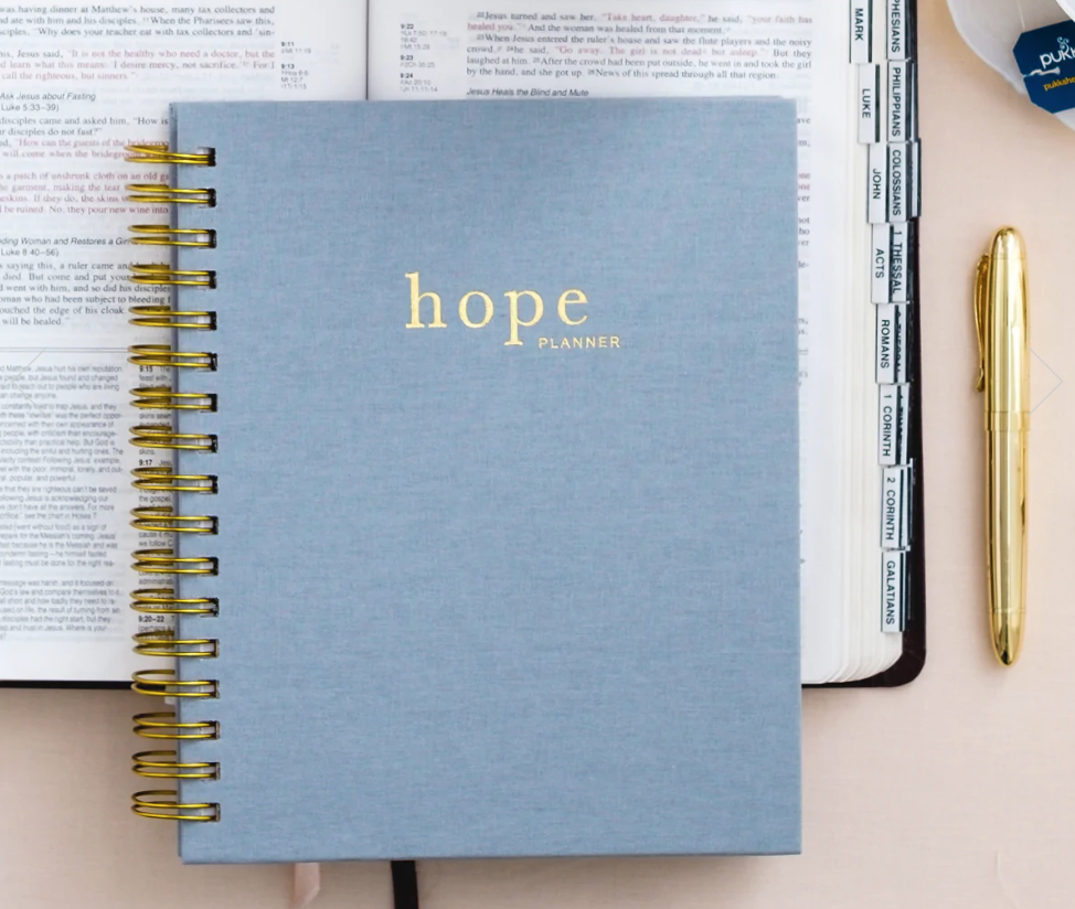 2023 Hope planner with blue spiral-bound cover sitting atop the Bible with a gold pen nearby