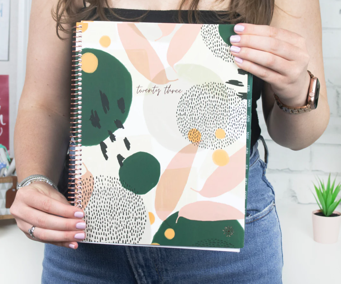 White woman in green tank top holding the Bloom 2023 daily planner in abstract green cover design