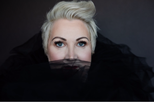 Kelly Diels, a white woman with white blonde short hair, wearing black tulle around her neck and her face peeking over the edge to reveal her nose and eyes.
