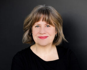 Headshot of a white woman with brown and gray hair, wearing a black top and red lipstick